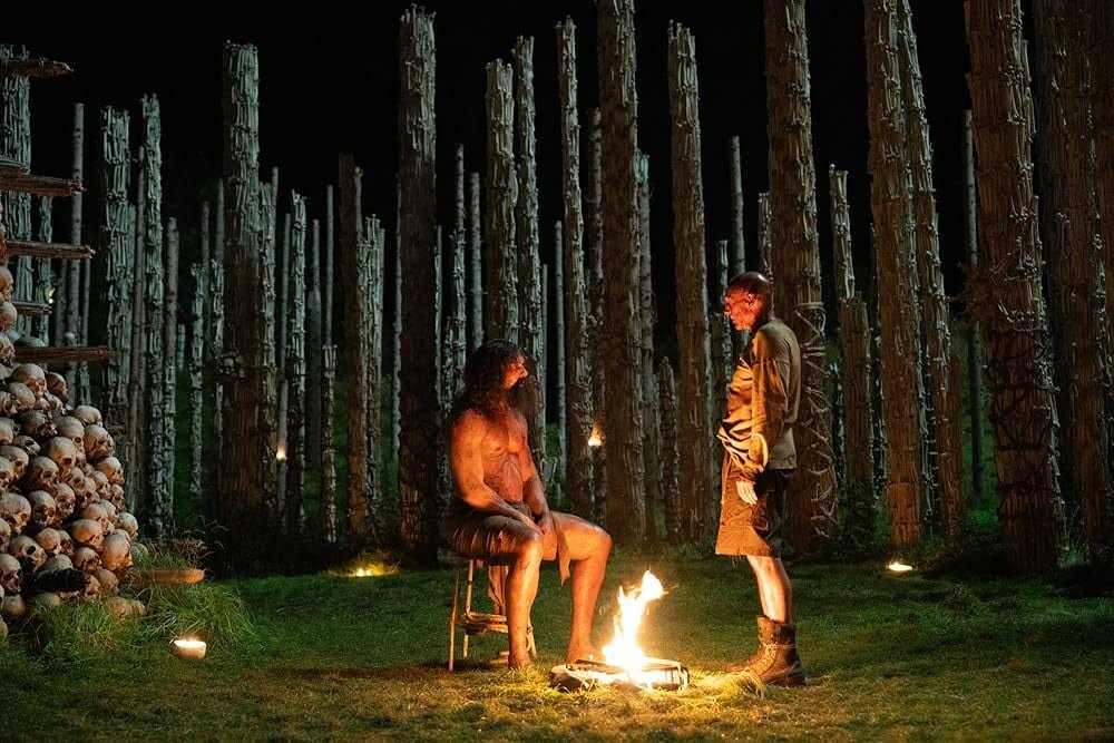 A seated, shirtless man is surrounded by a pile of pones and a large group of pillars. Next to him stands a man in dirty clothes.