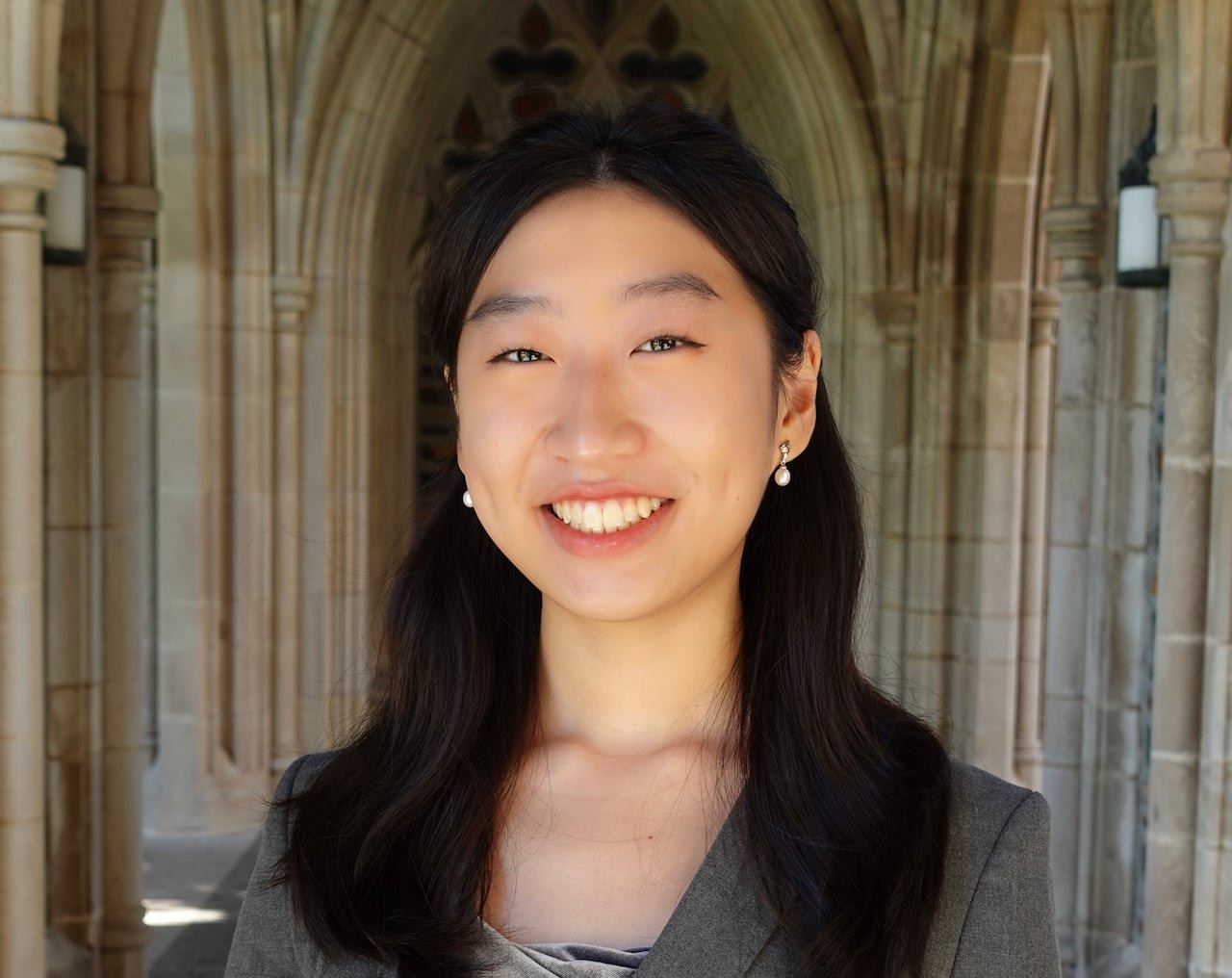 default-16 A smiling student with long dark hair and a grey blazer, standing in a stone alcove.