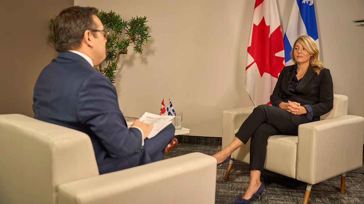 A woman in a suit sits in a chair in front of a Canadian and Quebec flag. A man in a suit sits across from her facing away from the camera.
