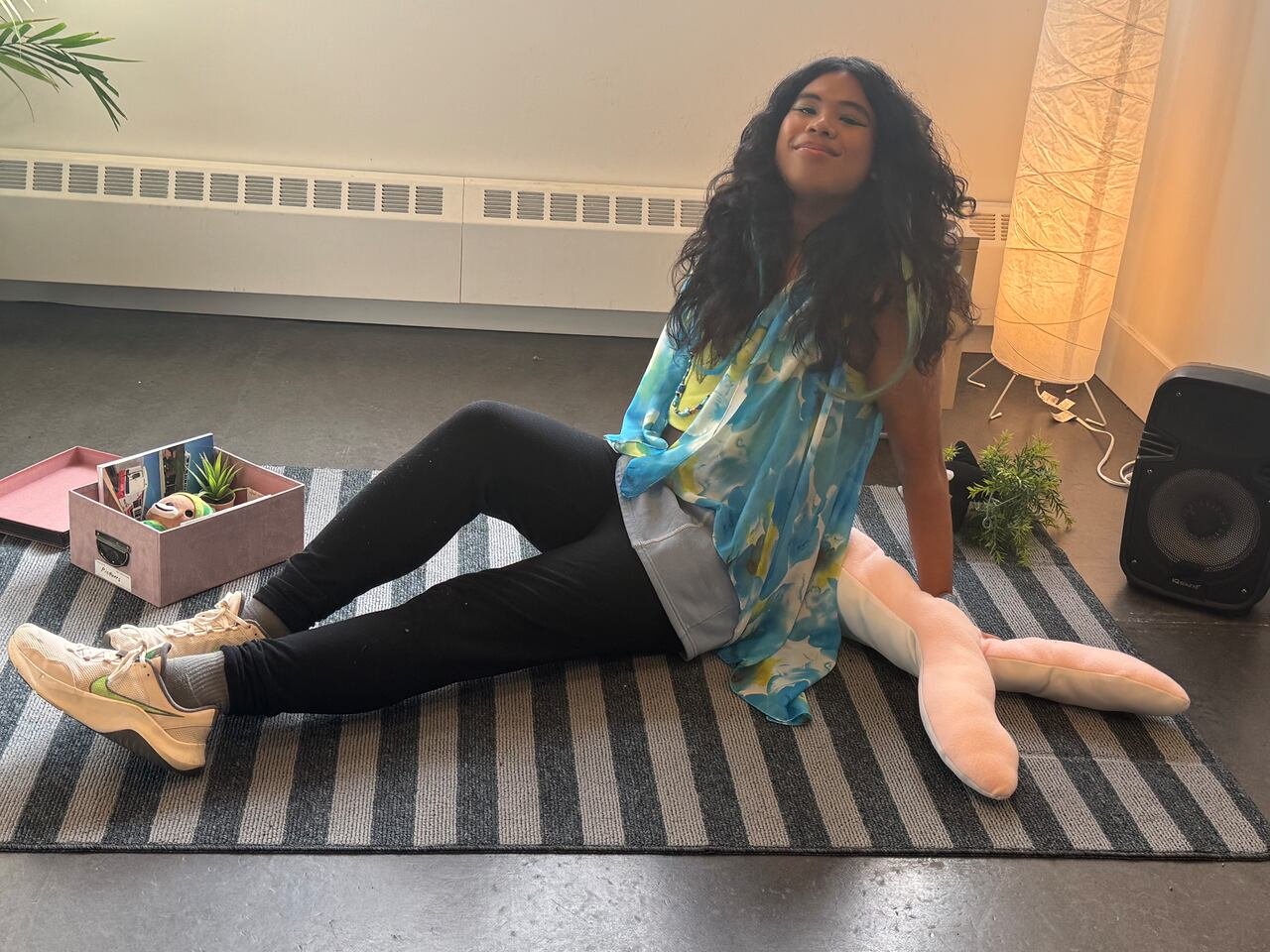 A queer youth with curly long dark hair sits on a carpet and smiles at the camera