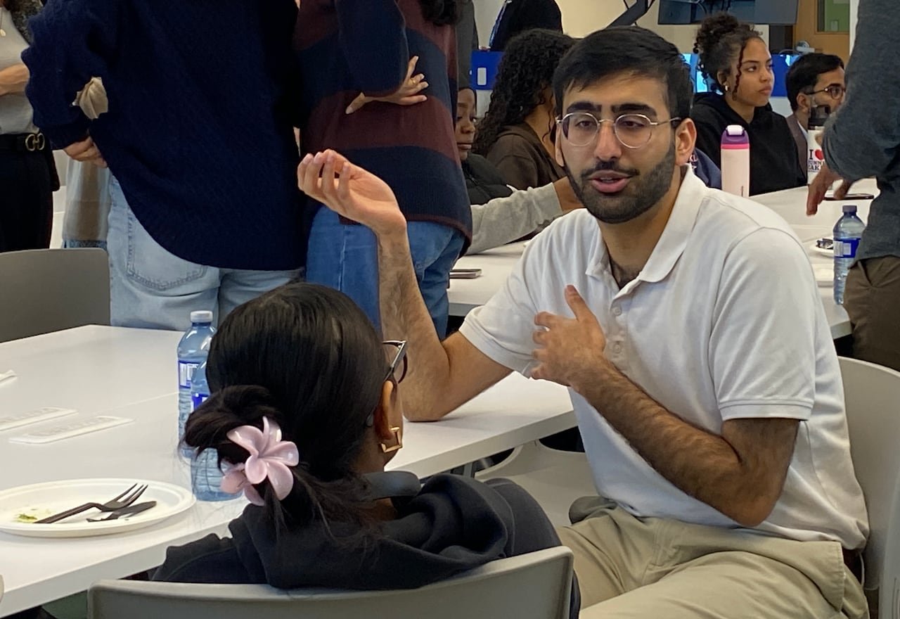 Students sit and stand around large conference tables in a bright room for discussions, as instructors walk around.