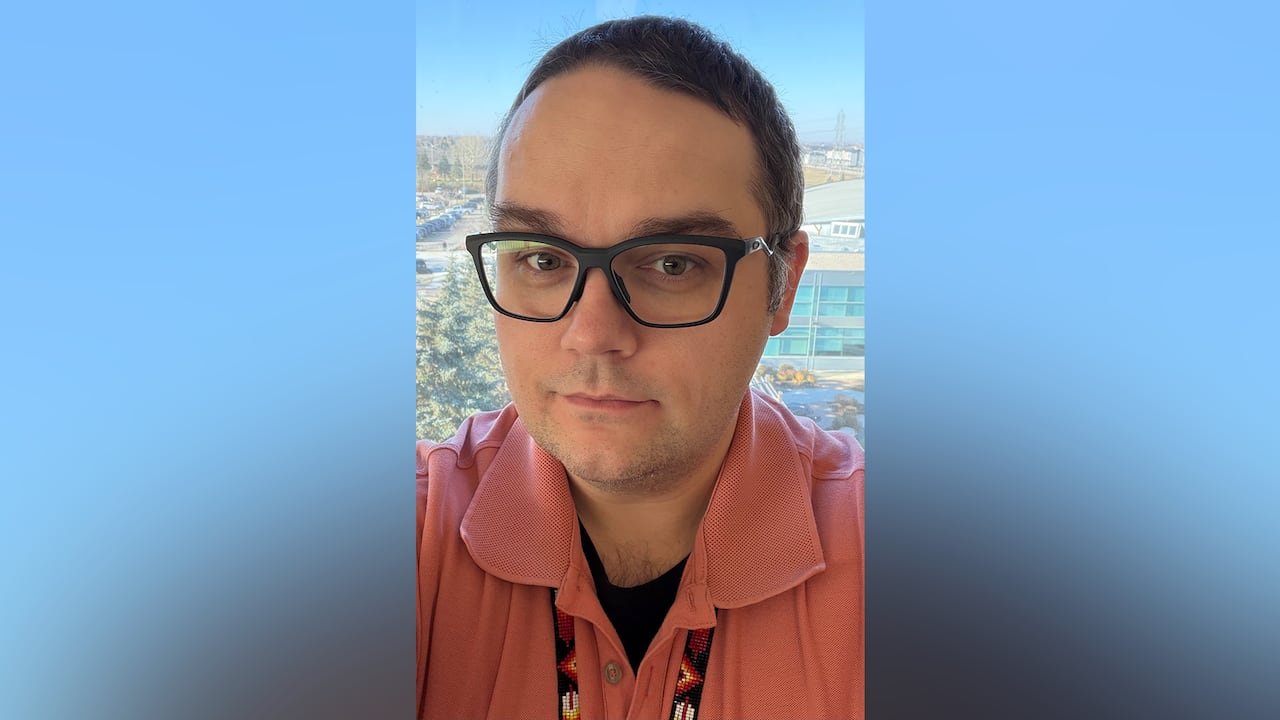 A portrait of a man wearing a coral polo shirt, glasses and a beaded lanyard.