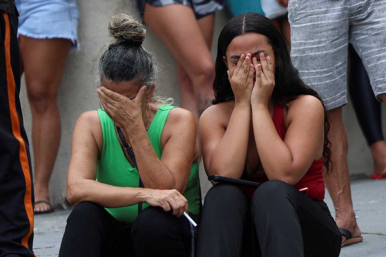 Two women are shown seated and wearing light clothing, their hands covering their faces in apparent distress.
