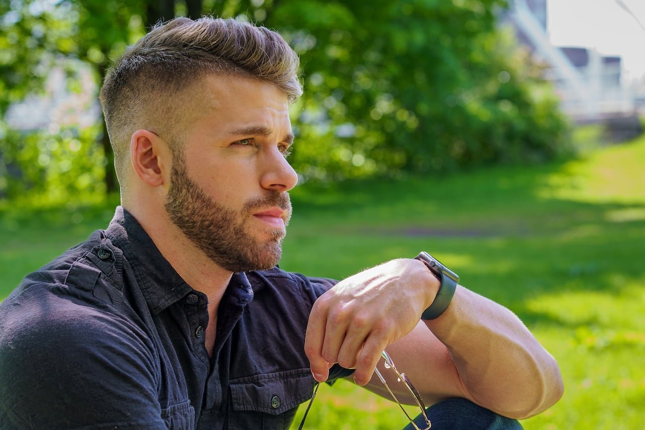 A man with brown hair and a dark shirt sits on a grassy field.